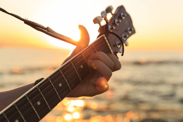 close up on the neck of a guitar with a person's hand playing a chord. ocean and sunset in the background